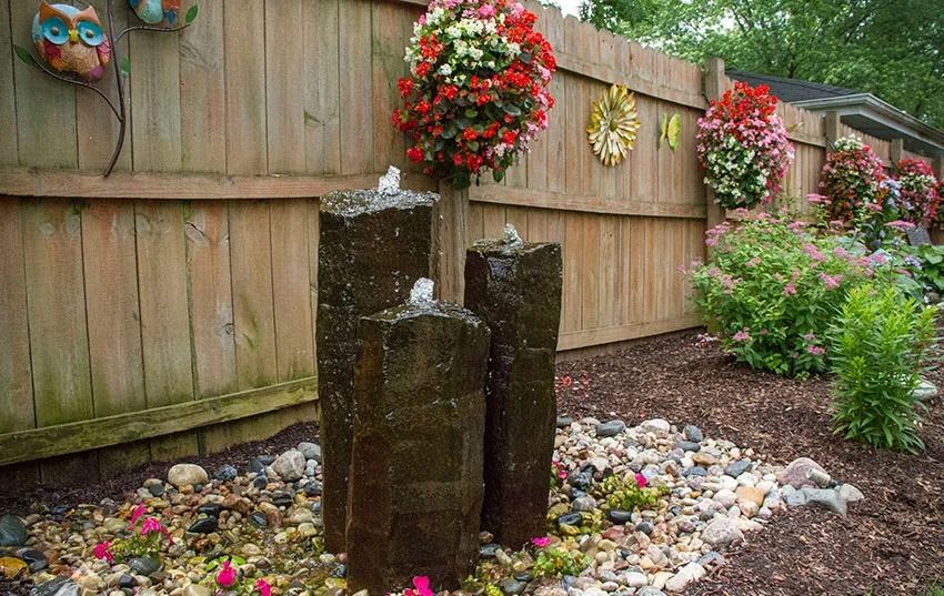 Three basalt column fountains set in river rock beside a wood fence and flower beds